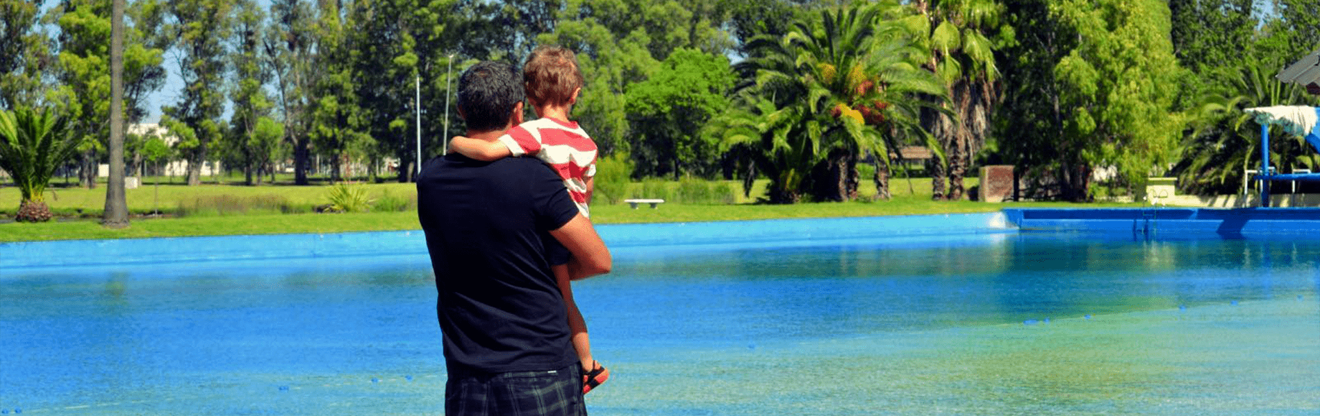 Hombre adulto con un niño en brazos miran la pileta, estan de espalda.